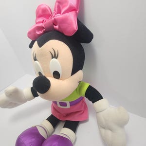 May include: A plush Minnie Mouse doll with a large pink bow, wearing a green top, pink skirt, and purple shoes. The doll has black ears, a beige face, and white gloves. The doll is sitting.