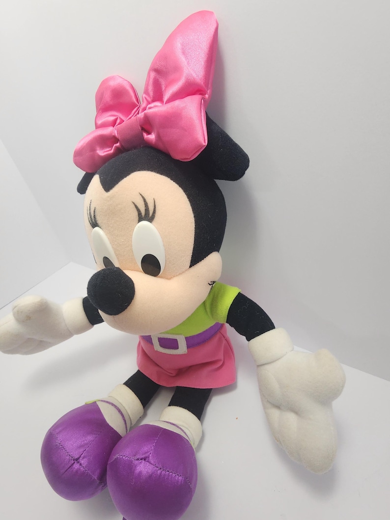 May include: A plush Minnie Mouse doll with a large pink bow, wearing a green top, pink skirt, and purple shoes. The doll has black ears, a beige face, and white gloves. The doll is sitting.