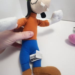 May include: A plush toy of Goofy from the Disney cartoon series. He is wearing a green hat, an orange shirt, blue trousers, and brown shoes. The toy has a SEGA tag attached to its trousers.