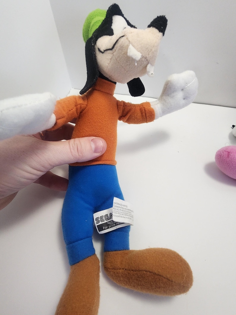 May include: A plush toy of Goofy from the Disney cartoon series. He is wearing a green hat, an orange shirt, blue trousers, and brown shoes. The toy has a SEGA tag attached to its trousers.