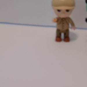 Puede incluir: Una peque&ntilde;a figura de juguete de pl&aacute;stico de una persona que lleva un sombrero marr&oacute;n y una camisa marr&oacute;n con pantalones marrones. La figura est&aacute; de pie sobre una superficie blanca.