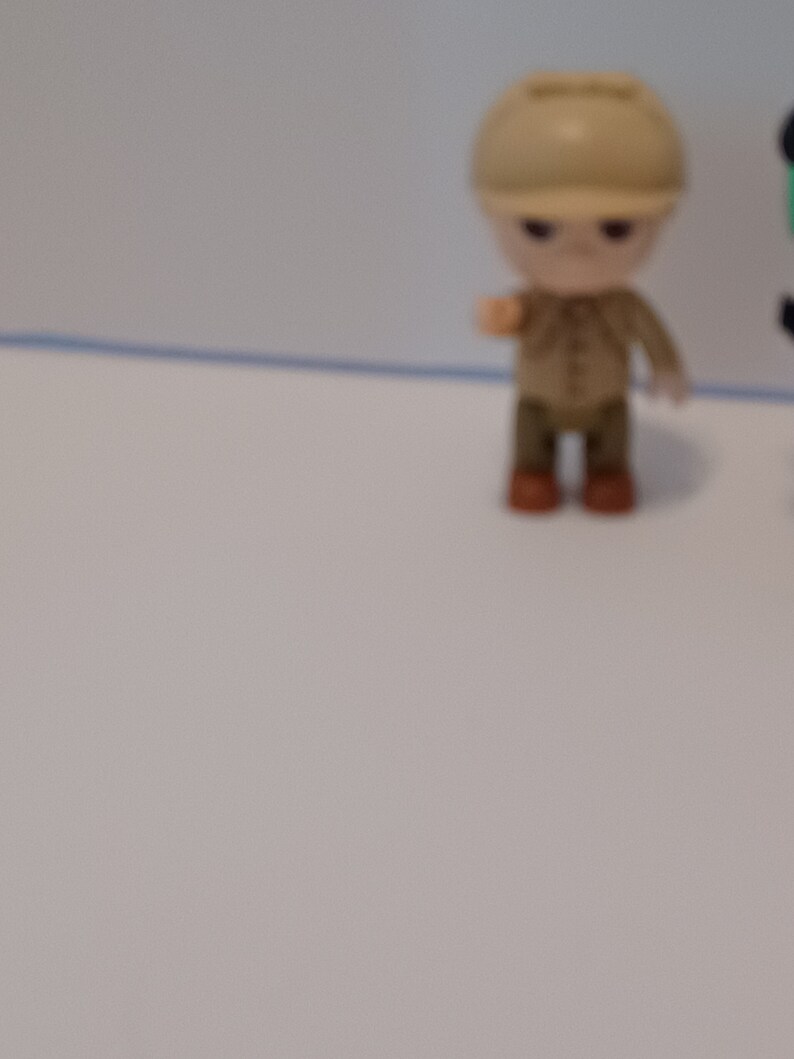 Puede incluir: Una peque&ntilde;a figura de juguete de pl&aacute;stico de una persona que lleva un sombrero marr&oacute;n y una camisa marr&oacute;n con pantalones marrones. La figura est&aacute; de pie sobre una superficie blanca.