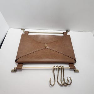 May include: A brown faux leather wall-mounted organizer featuring a central envelope-style pocket, two horizontal brass-toned metal rods, and four removable brass-toned S-hooks hanging from the bottom rod for storage and organization.