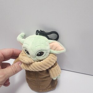 May include: A plush Grogu keychain featuring a light green head, large black eyes, and tan ears. The figure is wearing a brown coat and a tan collar. A black clip is attached to the top.