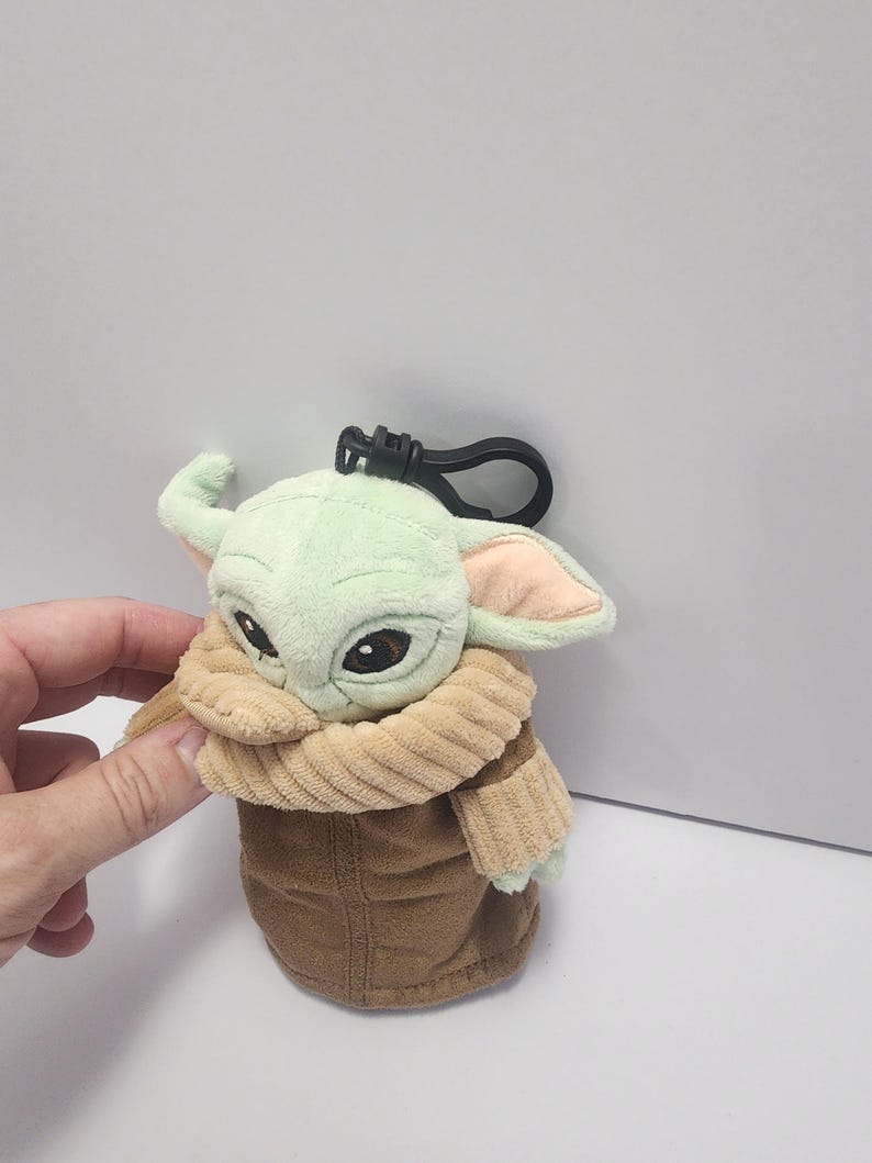 May include: A plush Grogu keychain featuring a light green head, large black eyes, and tan ears. The figure is wearing a brown coat and a tan collar. A black clip is attached to the top.