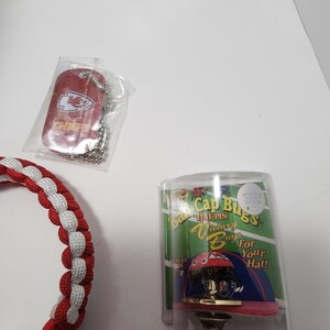 May include: A red dog tag with the Kansas City Chiefs logo and text, a red and white braided bracelet, and a hat pin in a clear plastic case. The hat pin is a miniature football helmet.