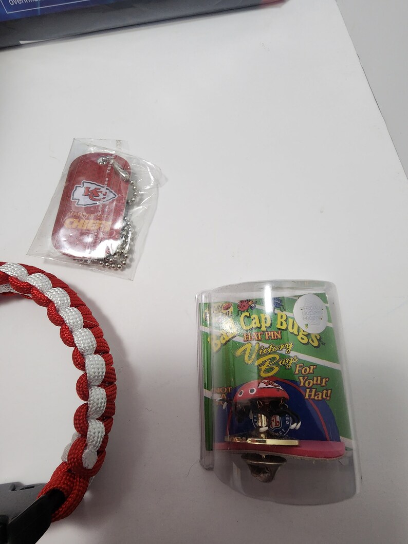 May include: A red dog tag with the Kansas City Chiefs logo and text, a red and white braided bracelet, and a hat pin in a clear plastic case. The hat pin is a miniature football helmet.