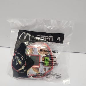 May include: A sealed plastic bag containing a McDonald's Happy Meal toy. The toy is a football-themed electronic game featuring Michael Vick. The packaging includes the ESPN logo and text in English, French, and Spanish.