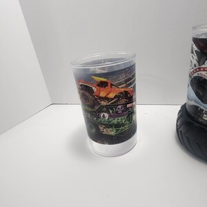 May include: A clear plastic tumbler featuring a colorful monster truck design. The design includes an orange truck with a yellow surfboard, a green truck, and a stadium background. The tumbler has a white base and a clear upper section.