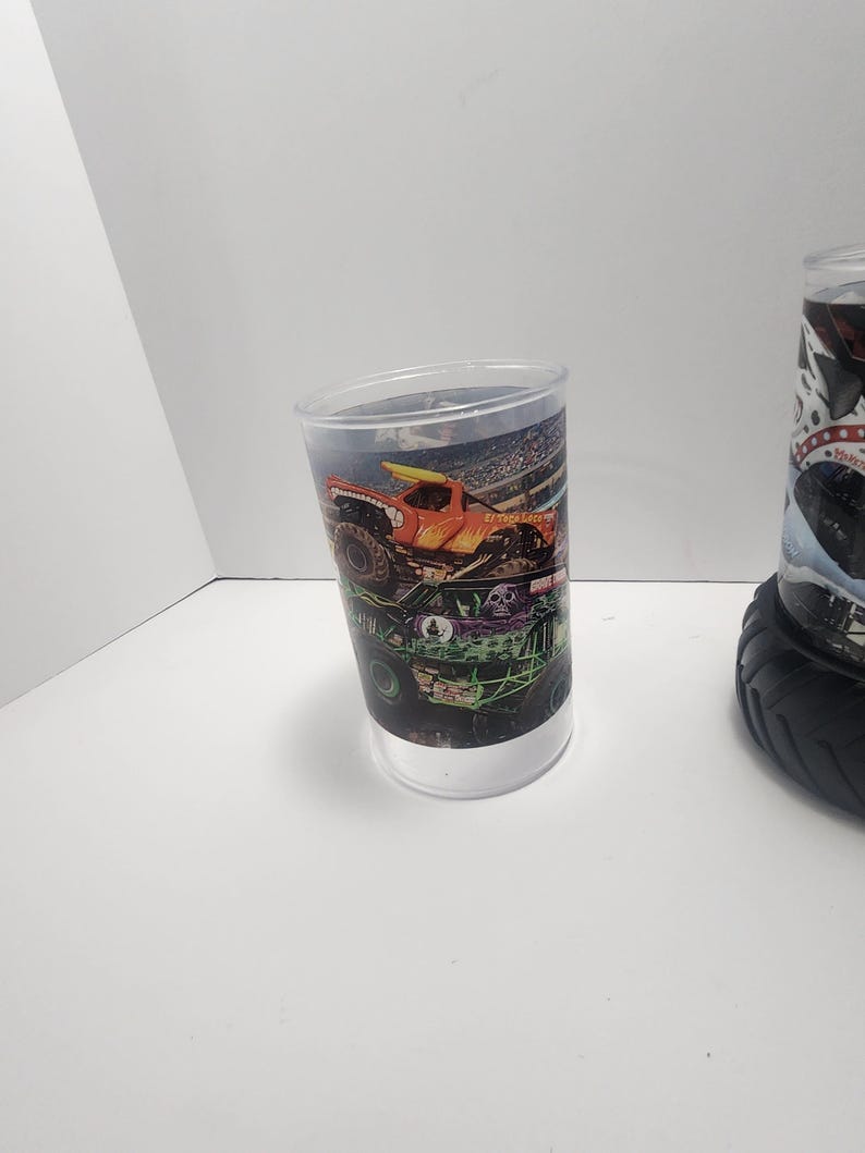 May include: A clear plastic tumbler featuring a colorful monster truck design. The design includes an orange truck with a yellow surfboard, a green truck, and a stadium background. The tumbler has a white base and a clear upper section.
