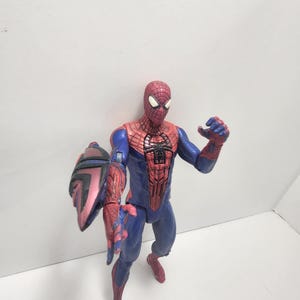 May include: A Spider-Man action figure in a dynamic pose, wearing a red and blue costume with web detailing. The figure has a shield attached to its left arm. The figure is standing on a white surface.