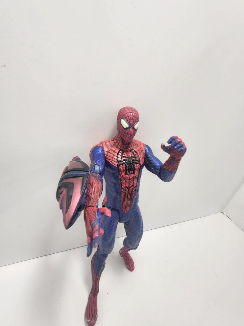 May include: A Spider-Man action figure in a dynamic pose, wearing a red and blue costume with web detailing. The figure has a shield attached to its left arm. The figure is standing on a white surface.