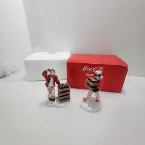 May include: Two ceramic Coca-Cola figurines depicting delivery workers in red jackets and red-and-white striped trousers. One figure drinks from a bottle beside a stack of crates, while the other carries a crate. Includes a red Coca-Cola box.