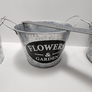 Może przedstawiać: Ocynkowane metalowe wiadro z czarną owalną etykietą z białym napisem "FLOWERS & GARDEN". Wiadro ma srebrny uchwyt i średnicę około 15 cm. Na górze wiadra umieszczono linijkę.