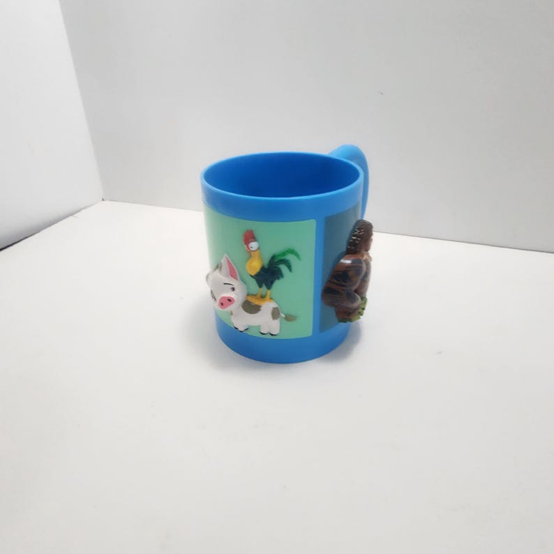 May include: A blue plastic mug with a handle. The mug features a light green panel with cartoon images of a pig, rooster, and cow. A brown figure is on the side of the mug.