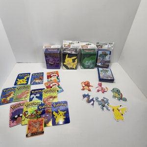 May include: A collection of Pokemon trading cards and stickers featuring various characters, including Pikachu, Charmander, Squirtle, and Bulbasaur. The cards are holographic and have a variety of colours, including blue, red, yellow, and green. The stickers are also holographic and feature the same characters. The cards and stickers are all in excellent condition.