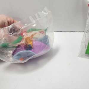 May include: A clear plastic bag containing colourful plastic toys. The toys include blue, purple, and orange shapes, along with green and red pieces. The bag is held by a hand.