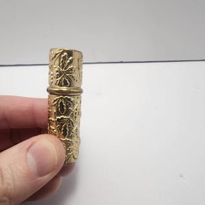 May include: A gold-colored, cylindrical perfume bottle with a leaf pattern. The bottle is held in a hand and has a hinged top. The design is embossed with a repeating leaf pattern. The bottle is approximately 3 inches tall.