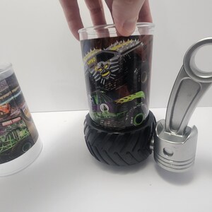 May include: A clear glass with a monster truck design, resting on a black tire-shaped base. The glass features a black and green monster truck graphic with the text "MAX-D". A silver piston is next to the glass.