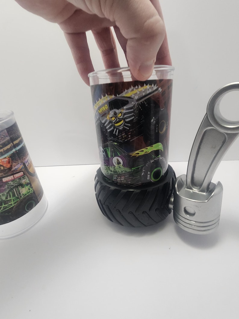 May include: A clear glass with a monster truck design, resting on a black tire-shaped base. The glass features a black and green monster truck graphic with the text "MAX-D". A silver piston is next to the glass.