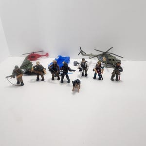 May include: A set of military action figures with two helicopters, a dog, and a blue tarpaulin. The figures are wearing camouflage uniforms and carrying weapons. One helicopter has the word "Police" printed on the side.