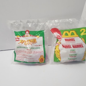 May include: Two sealed Happy Meal toys. One features Spider-Man with "Hobgoblin Landglider Vehicle" text. The other features Marvel Superheroes with "Storm Vehicle" text. Both toys are in clear plastic bags with green and yellow accents.