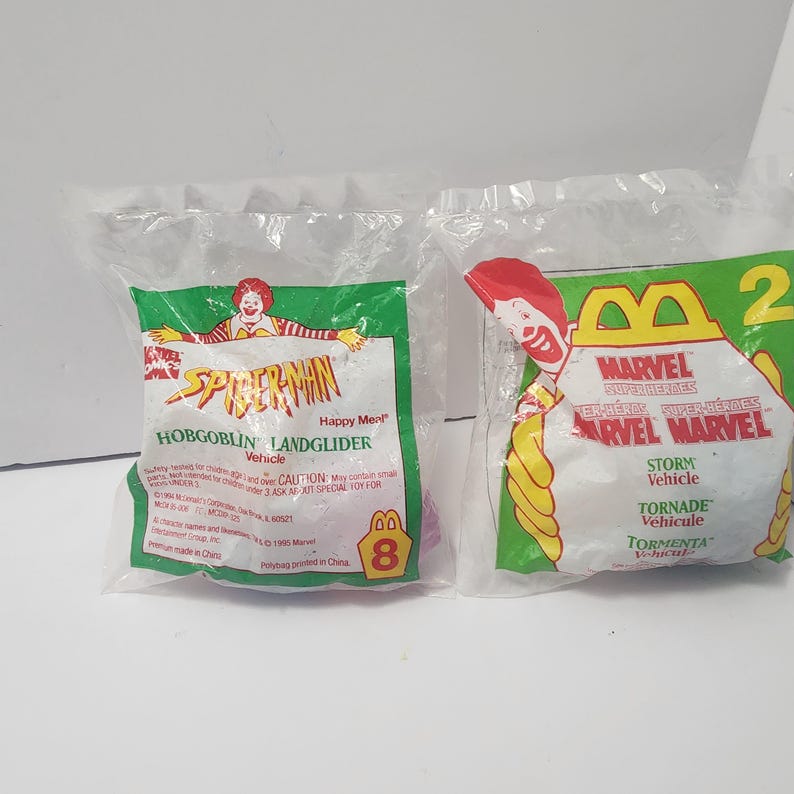May include: Two sealed Happy Meal toys. One features Spider-Man with "Hobgoblin Landglider Vehicle" text. The other features Marvel Superheroes with "Storm Vehicle" text. Both toys are in clear plastic bags with green and yellow accents.