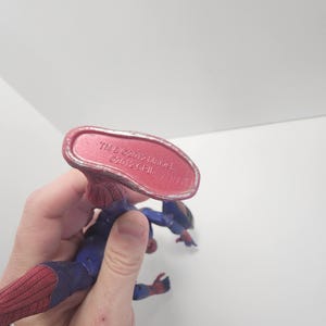 May include: A Spider-Man action figure in a dynamic pose, with a red and blue costume and web detailing. The figure's underside is visible, with text that reads "TM & &copy; 2012 Marvel". The figure is held against a white background.