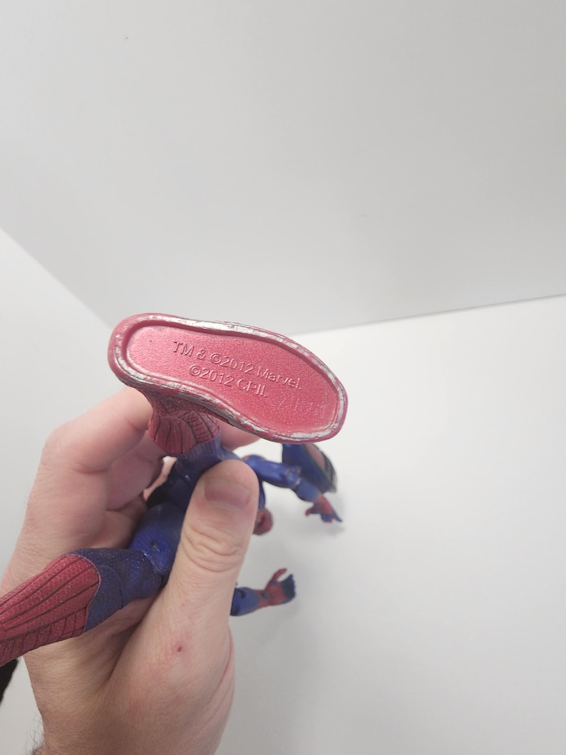 May include: A Spider-Man action figure in a dynamic pose, with a red and blue costume and web detailing. The figure's underside is visible, with text that reads "TM & &copy; 2012 Marvel". The figure is held against a white background.