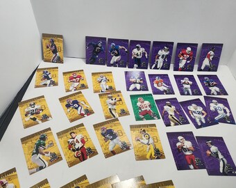 Könnte beinhalten: Eine Sammlung von Football-Sammelkarten mit verschiedenen Spielern in Action-Posen. Die Karten haben ein Gold- und Lila-Farbschema mit dem Text "First & Ten". Die Karten sind auf einer weißen Oberfläche angeordnet.