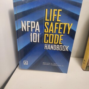 Peut inclure: Un livre à couverture rigide intitulé "NFPA 101 Life Safety Code Handbook" avec une couverture bleue et blanche. Le livre présente le logo NFPA et les noms des auteurs. Le livre est sur une surface blanche.