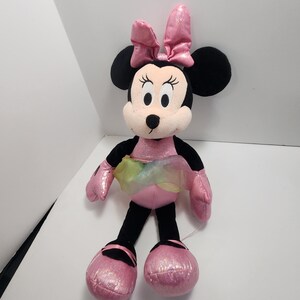 May include: A plush Minnie Mouse doll with a pink bow, dress, and shoes. The doll has black ears, arms, and legs. A rainbow-coloured tulle skirt adds a touch of whimsy. The doll is approximately 30 cm tall.