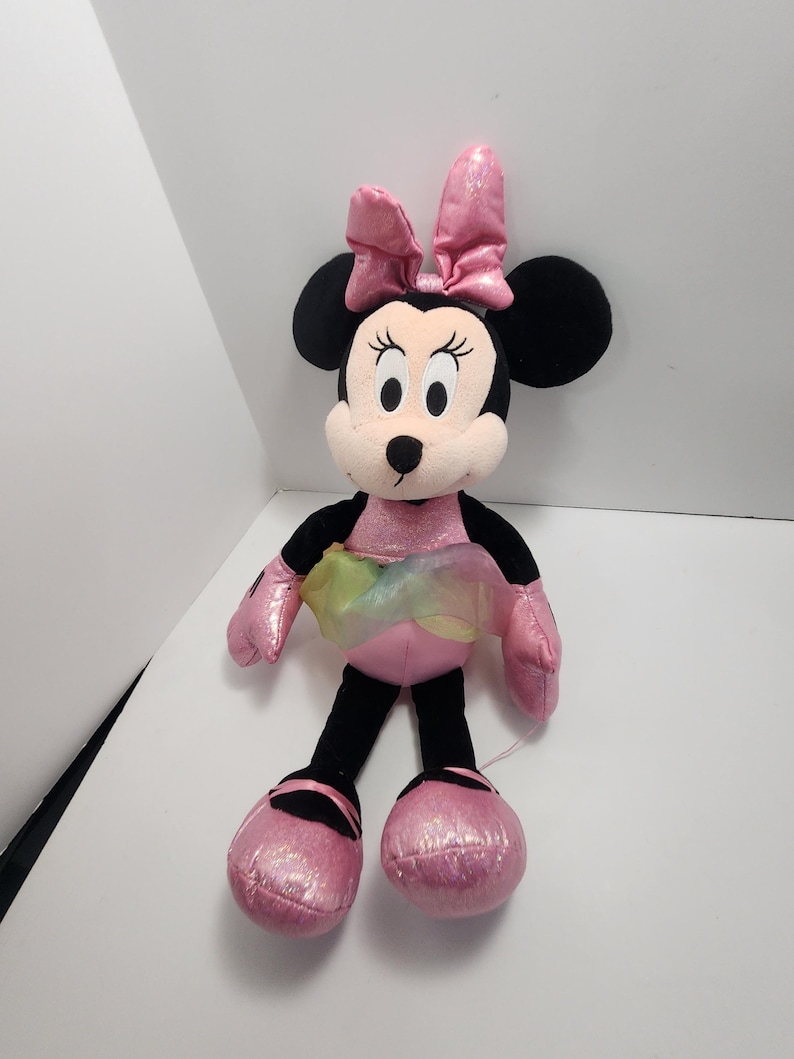 May include: A plush Minnie Mouse doll with a pink bow, dress, and shoes. The doll has black ears, arms, and legs. A rainbow-coloured tulle skirt adds a touch of whimsy. The doll is approximately 30 cm tall.