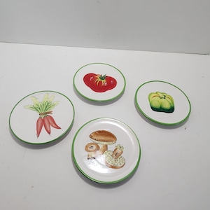 May include: Four round white ceramic plates with a green rim, each featuring a different hand-painted vegetable or fungi illustration: a bunch of red radishes, a red tomato, a green bell pepper, and a cluster of brown mushrooms.