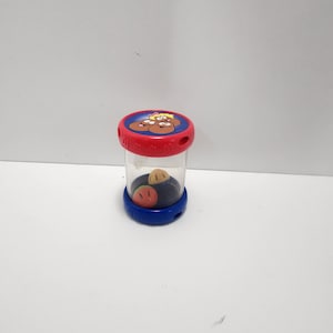 May include: A clear plastic toy with a red top and blue base. The top features a cartoon design on a blue background. Inside the container are three small, colorful shapes. The toy is designed for children.
