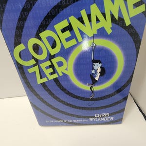 Könnte beinhalten: Ein Buch mit dem Titel "Codename Zero" mit einem blau-schwarzen Hintergrund mit Binärcode-Mustern. Der Titel ist in großen, grünen Buchstaben gehalten. Eine Cartoonfigur hängt an einem Seil in einem grünen Kreis. Der Autor ist Chris Rylander.