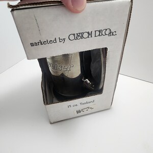 Puede incluir: Una caja blanca con el texto "marketed by CUSTOM DEC Inc." y "15 oz. Tankard" impreso en ella. La caja contiene un tanque de plata con la palabra "weiser" en relieve.