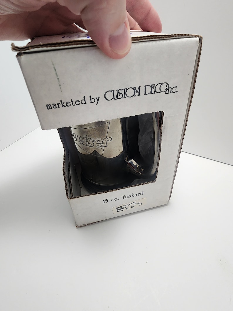 Puede incluir: Una caja blanca con el texto "marketed by CUSTOM DEC Inc." y "15 oz. Tankard" impreso en ella. La caja contiene un tanque de plata con la palabra "weiser" en relieve.