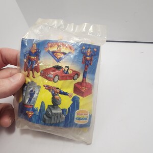 May include: Sealed plastic bag with Superman-themed toys. The toys include Superman figures, a red car, and a cityscape base. The packaging features the Superman logo and the Burger King Kids Club logo.