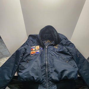Può includere: Giacca bomber blu navy con colletto in finta pelliccia nera e diverse toppe. La giacca ha orlo e polsini neri a coste e chiusura con cerniera. Le toppe includono bandiere statali e altri emblemi.