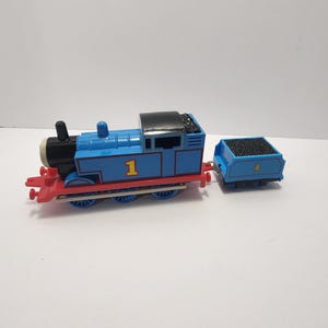 May include: A blue toy train with the number 1 on the side, a black roof, and a red front. It is connected to a blue coal car with the number 4. The toy is on a white surface.