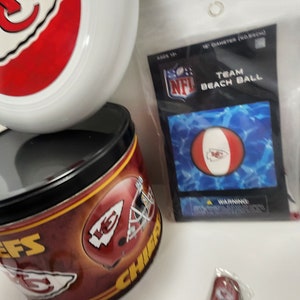 May include: A collection of Kansas City Chiefs merchandise, including a white and red frisbee, a tin with team graphics, and a beach ball in a package. The beach ball is 18 inches in diameter.