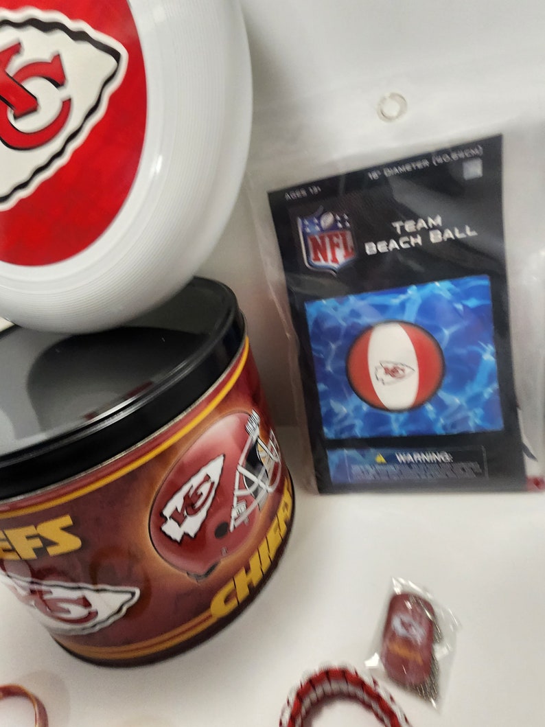 May include: A collection of Kansas City Chiefs merchandise, including a white and red frisbee, a tin with team graphics, and a beach ball in a package. The beach ball is 18 inches in diameter.