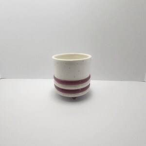 May include: A small, off-white ceramic planter with a speckled finish. The planter features two horizontal burgundy stripes around the middle and three small feet. The interior is a solid off-white color.