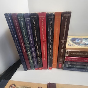 May include: A collection of Lemony Snicket's "A Series of Unfortunate Events" books. The spines are various colors, including blue, red, and brown. The books are arranged on a white surface, with some stacked and others standing upright.