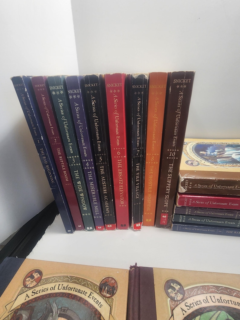 May include: A collection of Lemony Snicket's "A Series of Unfortunate Events" books. The spines are various colors, including blue, red, and brown. The books are arranged on a white surface, with some stacked and others standing upright.