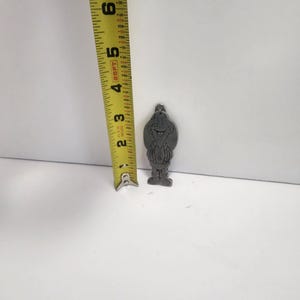 May include: A small, silver-colored metal charm in the shape of a cartoon character, approximately 2 inches tall. The charm features a smiling face and a detailed outfit. A yellow measuring tape is next to the charm.