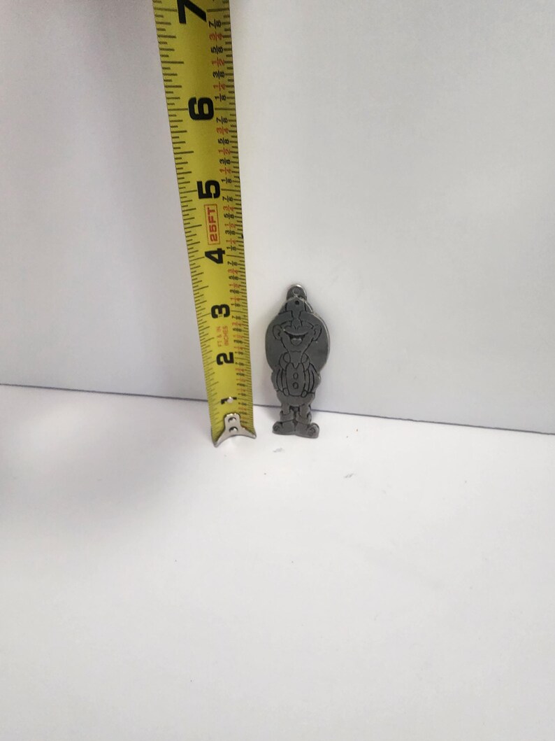 May include: A small, silver-colored metal charm in the shape of a cartoon character, approximately 2 inches tall. The charm features a smiling face and a detailed outfit. A yellow measuring tape is next to the charm.