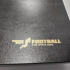 Peut inclure: Une bo&icirc;te noire avec un motif en relief dor&eacute;. La bo&icirc;te pr&eacute;sente une illustration stylis&eacute;e d'un joueur de football courant avec le ballon. Le texte "Thinking Man's Football A 3M Sports Game" est imprim&eacute; en or.