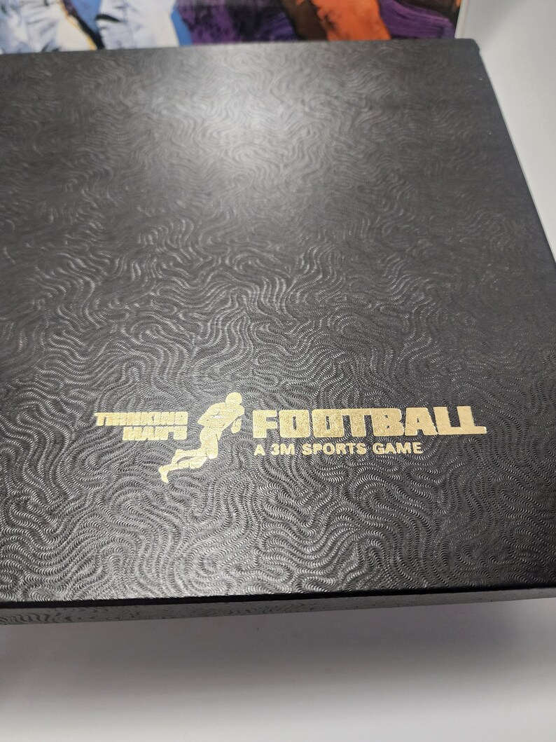 Peut inclure: Une bo&icirc;te noire avec un motif en relief dor&eacute;. La bo&icirc;te pr&eacute;sente une illustration stylis&eacute;e d'un joueur de football courant avec le ballon. Le texte "Thinking Man's Football A 3M Sports Game" est imprim&eacute; en or.